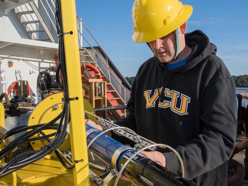 a v.c.u. student wearing a hard hat on a boat examines a piece of large equipment